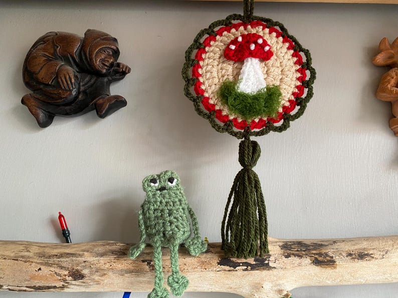 May include: A round crochet wall hanging featuring a red and white mushroom motif with a green base, surrounded by a red and dark green scalloped border and a long dark green tassel. Below it sits a small, light green crocheted frog figurine.