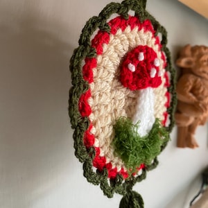 May include: A circular crochet wall hanging featuring a central beige disc with a red and white spotted mushroom motif. The mushroom is accented with green textured yarn at the base. The piece is framed by a red border and a dark green scalloped edge.