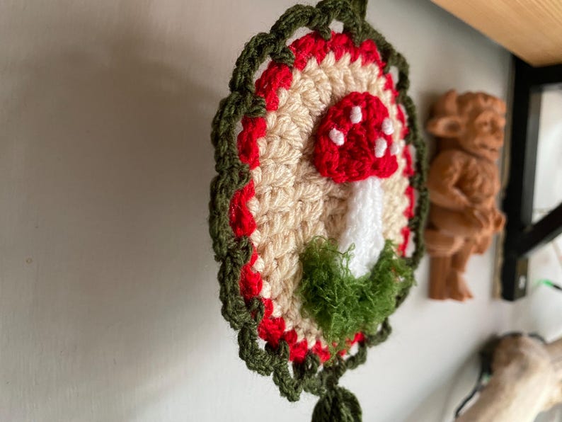 May include: A circular crochet wall hanging featuring a central beige disc with a red and white spotted mushroom motif. The mushroom is accented with green textured yarn at the base. The piece is framed by a red border and a dark green scalloped edge.