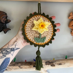 May include: A crocheted daffodil flower with a green tassel hanging from it. The flower is yellow and white with green leaves. The crocheted piece is hanging from a wooden surface.