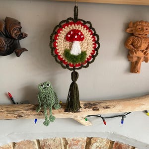 May include: A circular crochet wall hanging featuring a red and white mushroom motif on a beige background, framed by a dark green scalloped border and a matching tassel. Beside it sits a small, light green crocheted frog figure.
