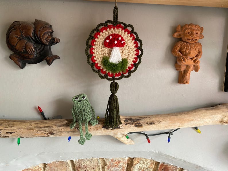 May include: A circular crochet wall hanging featuring a red and white mushroom motif on a beige background, framed by a dark green scalloped border and a matching tassel. Beside it sits a small, light green crocheted frog figure.