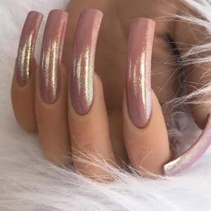 May include: Close-up of long, pink, iridescent acrylic nails. The nails have a glossy finish with a subtle shimmer effect. The nails are set against a backdrop of soft, white, fluffy material.