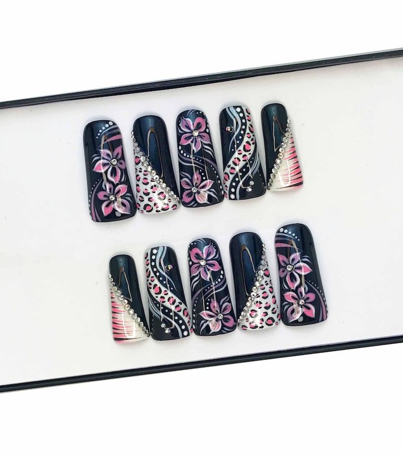 Puede incluir: Conjunto de diez u&ntilde;as postizas negras con dise&ntilde;os florales rosas y blancos y pedrer&iacute;a.