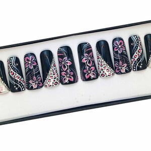 Puede incluir: Un conjunto de diez u&ntilde;as postizas negras con dise&ntilde;os florales rosas y blancos y pedrer&iacute;a.