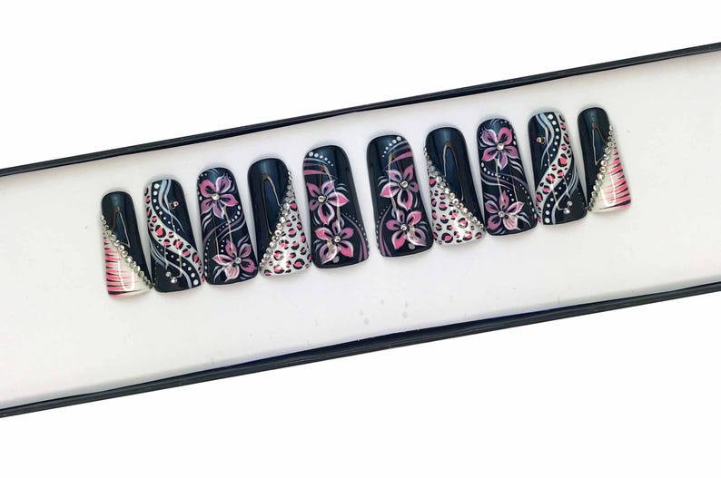 Puede incluir: Un conjunto de diez u&ntilde;as postizas negras con dise&ntilde;os florales rosas y blancos y pedrer&iacute;a.