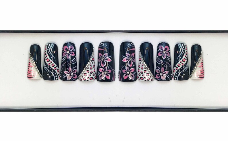 Puede incluir: Un conjunto de diez u&ntilde;as postizas negras con dise&ntilde;os florales rosas y blancos y pedrer&iacute;a.