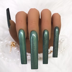 Op de afbeelding: Een set lange, vierkant gevormde kunstnagels met een metallic, glinsterende bosgroene afwerking. De nagels worden gepresenteerd op een neutraal gekleurd etalagepop-handmodel en tonen een glad, reflecterend oppervlak met fijne glitterdeeltjes.