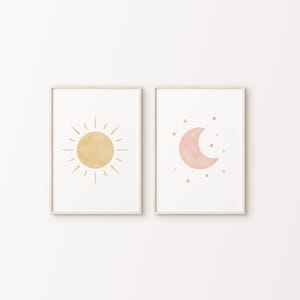 Pode incluir: Duas impressões emolduradas com ilustrações minimalistas de um sol amarelo e uma lua crescente rosa. A impressão do sol tem um fundo branco e a impressão da lua tem um fundo branco com pequenas estrelas douradas.