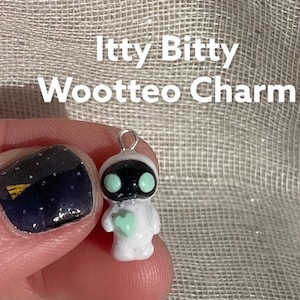 Könnte beinhalten: Ein kleiner, weißer Itty Bitty Wootteo Charm mit schwarzem Gesicht und mintgrünen Augen und Herz. Der Charm hat oben eine Silberschlaufe zum Befestigen an einer Halskette oder einem Armband. Der Text "Itty Bitty Wootteo Charm" ist weiß.