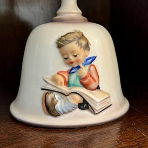 May include: A white porcelain bell with a hand-painted scene of a boy reading a book. The boy is wearing a red shirt, blue pants, and a blue and white scarf. The bell is decorated with gold trim.