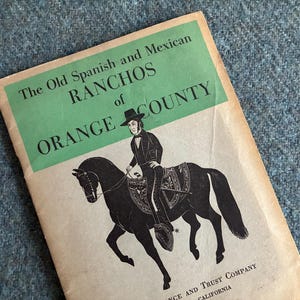 May include: Vintage book cover titled "The Old Spanish and Mexican Ranchos of Orange County." The cover features a black illustration of a person on horseback. The background is a textured blue surface.