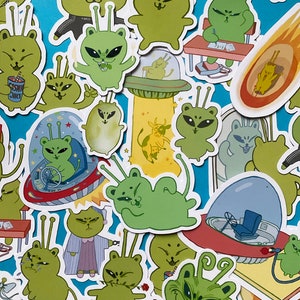 May include: A collection of colorful stickers featuring cartoon characters, including aliens, cats, and other whimsical creatures. The stickers are on a blue background and feature various designs and colors.