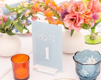 May include: A beautifully set table with floral arrangements, candles, and place settings. The centerpiece features a light blue table number card with the number 1. The table is decorated with colorful flowers, glassware, and blue napkins.