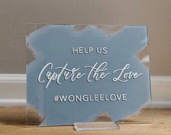 May include: Clear acrylic sign with a blue painted background and white script that reads "Help Us Capture the Love" and "#WONGLEELOVE". The sign is held up by a clear acrylic base.