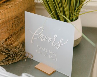 May include: A light blue sign with the word "Favors" in elegant white script. Below, the text reads "PLEASE TAKE ONE OF EACH." The sign is propped up by a small wooden stand, suggesting a wedding or event theme.