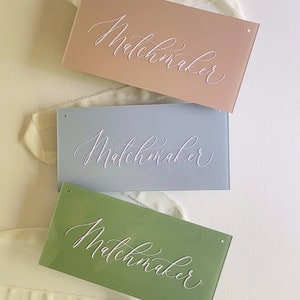 May include: Three rectangular signs in shades of peach, blue, and green, each with the word "Matchmaker" in elegant white script. The signs have small holes at the top and are arranged on a white surface with a ribbon.