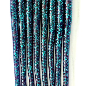 May include: A close-up shot of eight soft plastic fishing lures. The lures are a dark purple color with teal-colored speckles. The lures are cylindrical with a ribbed texture and tapered ends. The background is white.