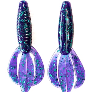 May include: Two purple and blue fishing lures with a speckled design. The lures have a ribbed, cylindrical body and a multi-lobed tail. The color scheme includes shades of purple, blue, and green, creating a vibrant and eye-catching appearance.