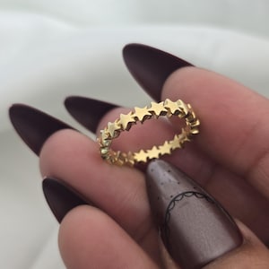Puede incluir: Un anillo dorado con un patrón continuo de estrellas. El anillo es de estilo banda, con cada estrella conectada a la siguiente, creando un círculo completo. El anillo se muestra sobre un fondo blanco suave.