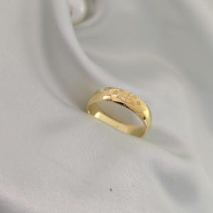 Peut inclure: Une bague en or avec des motifs gravés d'étoiles et de formes géométriques sur le bandeau. La bague est présentée sur un fond blanc texturé, soulignant sa surface polie et ses détails complexes. La couleur or chaud de la bague contraste avec le fond blanc.