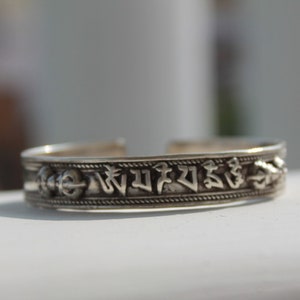 Puede incluir: Pulsera de plata con un diseño de rueda de oración tibetana. La pulsera está grabada con un mantra en escritura tibetana.