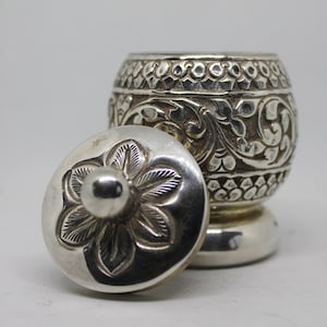May include: A silver container with a lid, both intricately engraved with floral patterns. The lid features a central flower design.
