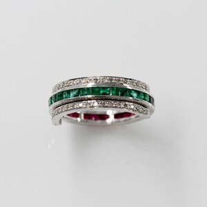 May include: A silver ring with three rows of gemstones. The top and bottom rows feature small, clear stones, while the middle row showcases vibrant green emeralds. The bottom row also has small, red gemstones.
