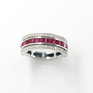 May include: A silver ring with three rows of gemstones. The middle row features square-cut, vibrant red rubies. The top and bottom rows are adorned with small, sparkling diamonds. The ring has a polished, reflective surface.