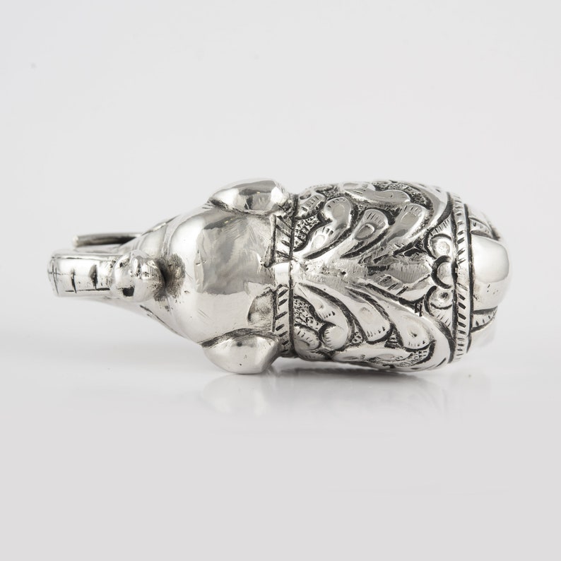 May include: A silver elephant figurine with intricate floral designs. The elephant is lying on its side with its trunk raised.