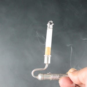 May include: A silver cigarette holder with a curved stem and a decorative design. A cigarette is inserted into the holder and is lit, with smoke rising from the end.