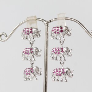 May include: Silver-toned elephant earrings with three dangling elephants per earring. Each elephant is adorned with small, round, pink gemstones. The earrings are displayed on a silver earring stand against a white background.