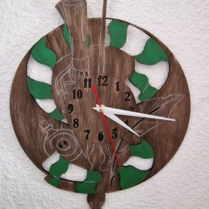 Puede incluir: Un reloj de madera con un diseño verde y blanco. La esfera del reloj tiene números romanos y las manecillas son rojas y blancas. El reloj tiene forma de círculo y tiene un péndulo decorativo.
