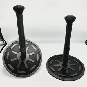 Op de afbeelding: Twee zwarte en grijze honkbal tees met een cirkelvormige basis en een verticale paal met een afgeronde knop. De basis heeft een gedetailleerd ontwerp. De tees staan tegen een witte achtergrond.