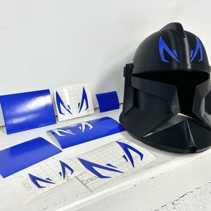 May include: A black helmet with blue decals, alongside blue vinyl sheets and decals. The helmet has blue markings on the forehead. The decals feature a stylized design. The items are arranged on a white surface.