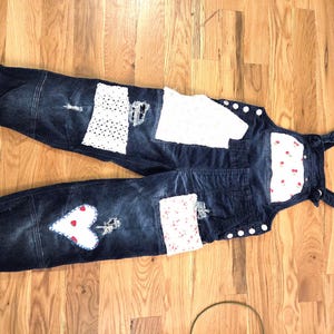 May include: Dark blue denim overalls with white patches and decorative stitching. Features a heart-shaped patch with red dots and a floral pattern on the cuffs. Includes a black hanger.