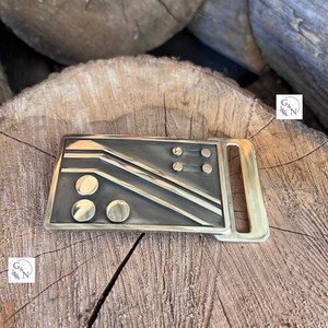 May include: A gold-toned metal belt buckle with a geometric design featuring black lines and circles. The buckle is on a wooden surface.
