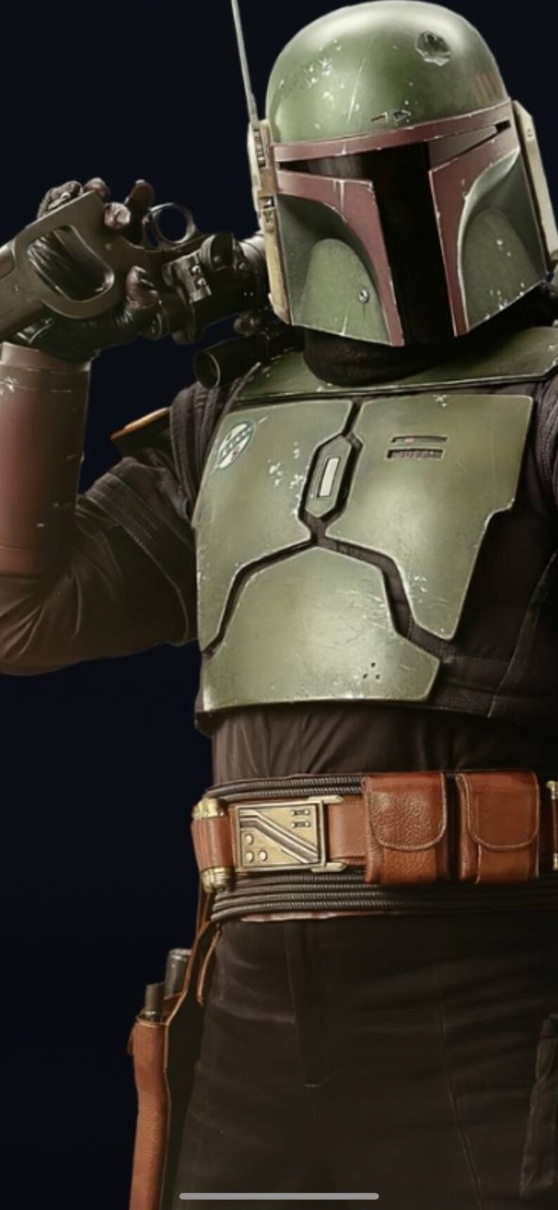 May include: A green and brown Mandalorian helmet with a visor. The helmet is worn by a person with a brown leather belt with a metal buckle. The belt has two pouches on the right side.