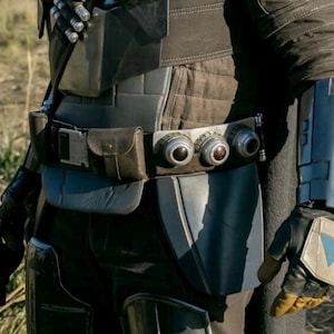 May include: Close-up of a costume featuring a dark gray and brown color scheme. The outfit includes a utility belt with three circular devices, a holster, and a forearm gauntlet with a blue accent. The gloves have a yellow palm and a blue triangle.