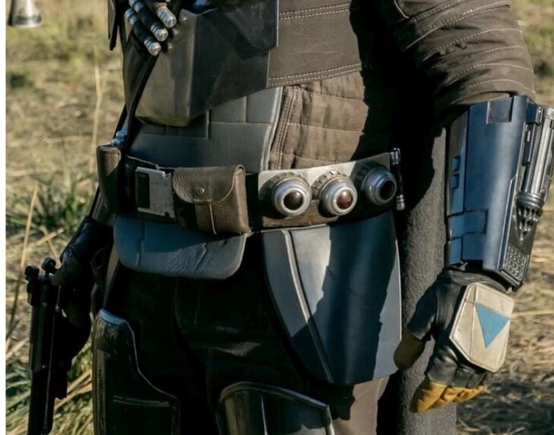 May include: Close-up of a costume featuring a dark gray and brown color scheme. The outfit includes a utility belt with three circular devices, a holster, and a forearm gauntlet with a blue accent. The gloves have a yellow palm and a blue triangle.