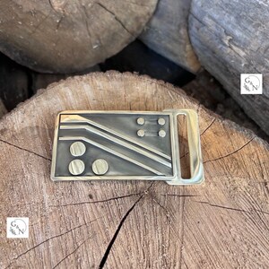May include: A gold-toned metal belt buckle with a geometric design featuring black lines and circles. The buckle has a smooth, polished finish.
