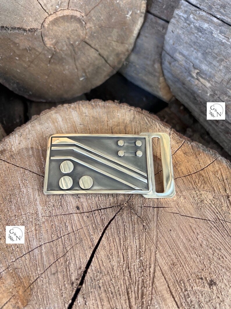 May include: A gold-toned metal belt buckle with a geometric design featuring black lines and circles. The buckle has a smooth, polished finish.