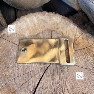 May include: A solid brass belt buckle with a rectangular shape and a single, centered, round prong. The buckle is sitting on a wooden surface.