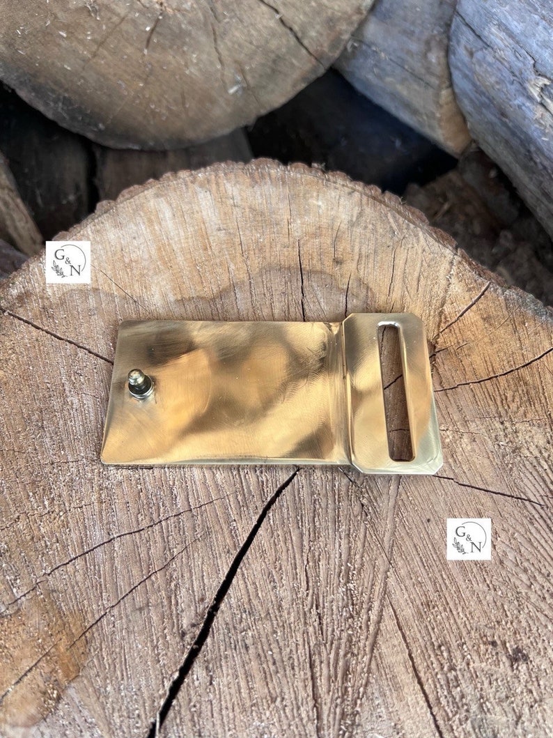 May include: A solid brass belt buckle with a rectangular shape and a single, centered, round prong. The buckle is sitting on a wooden surface.