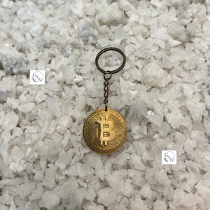Pode incluir: Um porta-chaves com moeda Bitcoin dourada. A moeda apresenta o símbolo Bitcoin e está presa a uma corrente e a um porta-chaves. O porta-chaves é exibido em um fundo branco texturizado.