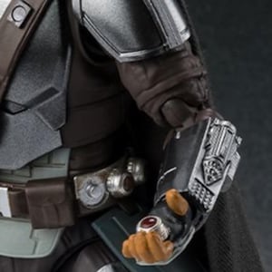 May include: Close-up of a Mandalorian action figure. The figure is clad in dark gray and silver armor, with a black cape. The figure's hand is visible, wearing a ring with a red stone. The figure is holding a silver weapon.