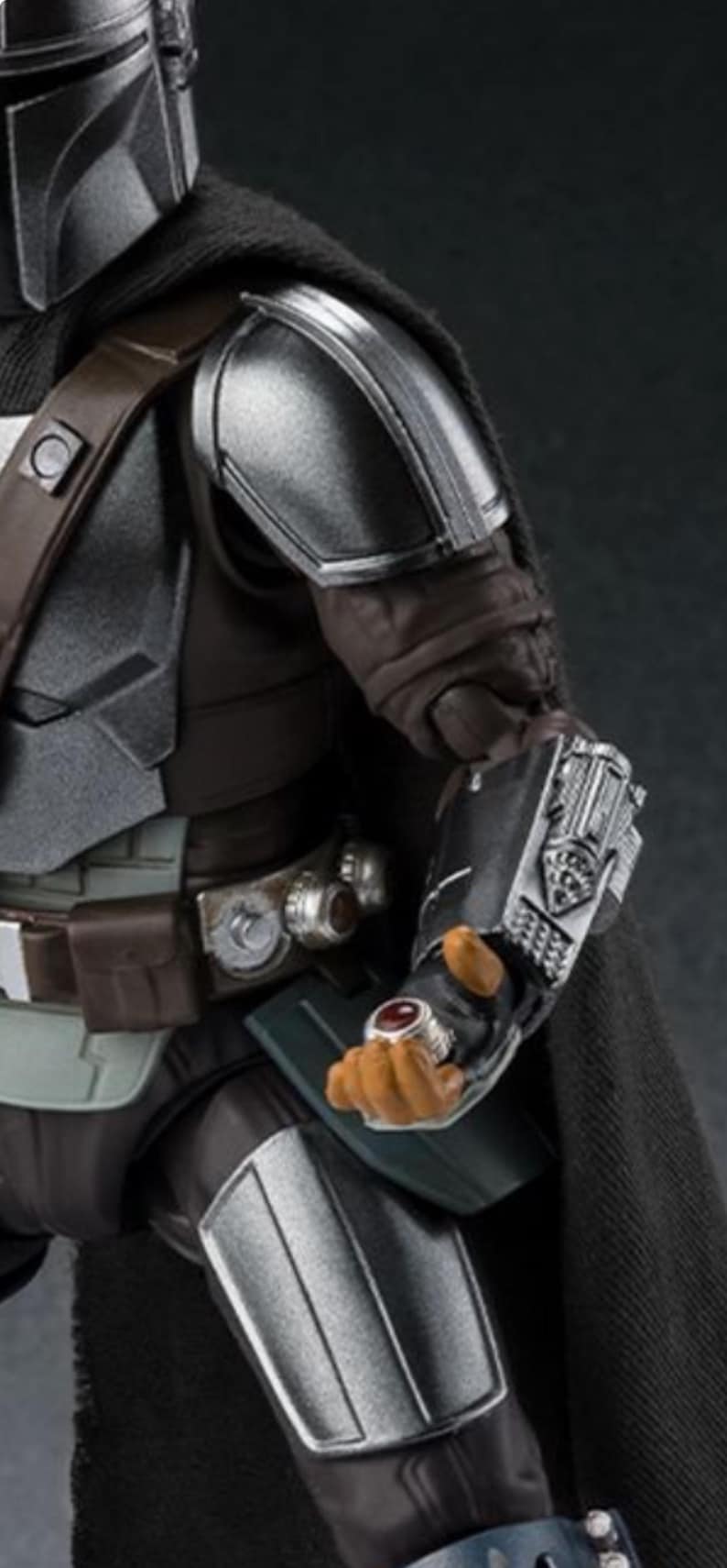 May include: Close-up of a Mandalorian action figure. The figure is clad in dark gray and silver armor, with a black cape. The figure's hand is visible, wearing a ring with a red stone. The figure is holding a silver weapon.