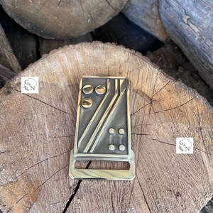 May include: A gold-toned metal belt buckle with a geometric design featuring black lines and circles. The buckle has a rectangular shape and a smooth, polished finish.