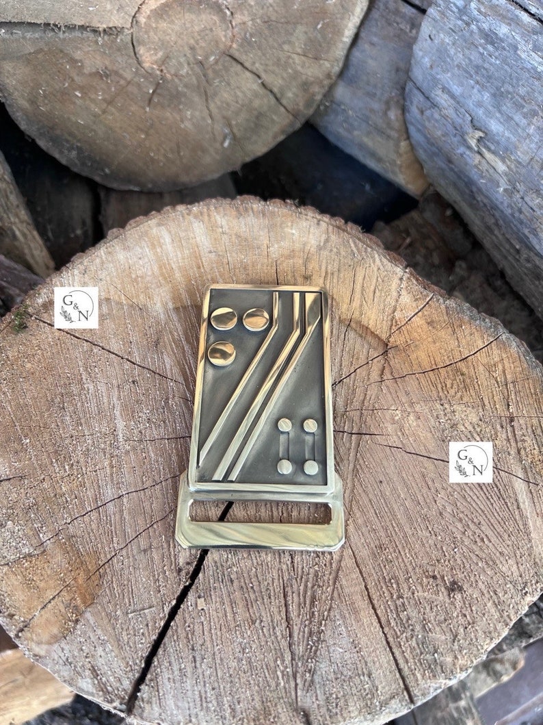 May include: A gold-toned metal belt buckle with a geometric design featuring black lines and circles. The buckle has a rectangular shape and a smooth, polished finish.