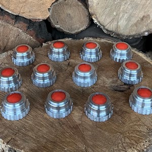 May include: A collection of twelve silver-colored bottle stoppers with a bright red, circular top. The stoppers are arranged on a wooden surface, showcasing their metallic finish and vibrant color contrast. The stoppers are approximately 1 inch in diameter.